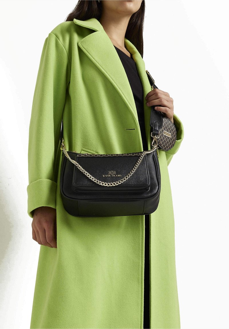 Borsa a tracolla in pelle nera con catena, caratterizzata da una superficie liscia e un logo impresso. Indossata insieme a un cappotto verde brillante.
