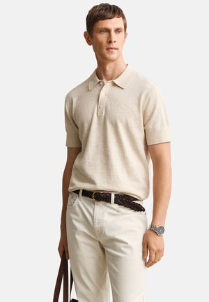 Homme portant un polo beige à manches courtes, pantalon clair avec une ceinture tressée foncée, montre, tenant la sangle d'un sac en cuir marron, regardant légèrement vers la droite.