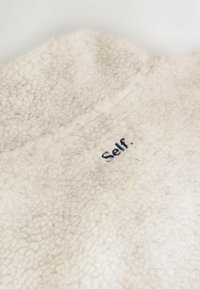 Crèmefarbener Fleece-Stoff mit einer strukturierten, flauschigen Oberfläche. An der Kante befindet sich ein kleiner, dunkelblauer, gestickter Text mit der Aufschrift "Selbst.".