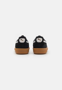 Scarpe sportive in suede nero con suola in gomma color miele, dotate di un logo bianco e oro sul tallone e accenti grigi lungo i lati.
