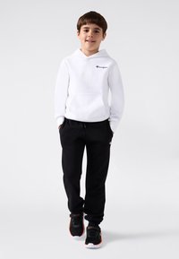 Junge steht in einem weißen Champion-Hoodie, schwarzen Jogginghosen und schwarzen Sneakers mit orangefarbenen und grünen Akzenten vor einem weißen Hintergrund.