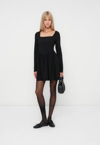 Vestido negro de manga larga con escote cuadrado y falda fruncida. Combinado con bailarina negra y un bolso negro texturizado. Medias negras transparentes.