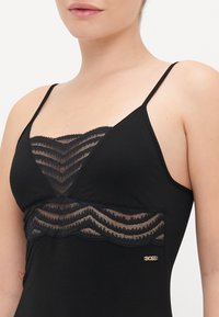 Svart cami med spetsinsats med zigzag-mönster, tunna justerbara axelband och en liten guldig logotyp på sidan.