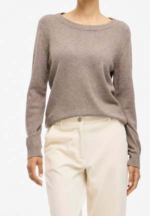 Frau trägt taupefarbenen, langärmeligen Strickpullover, der in hoch taillierte, elfenbeinfarbene Cordhosen gesteckt ist, steht vor einem schlichten Hintergrund.