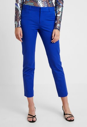 Femme portant un pantalon bleu ajusté longueur cheville, un top transparent à manches longues avec motif papillon et des sandales noires à talons ouvertes au niveau des orteils.