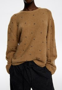 Suéter marrón de felpa con adornos de cuentas plateadas, escote ancho y ajuste holgado, combinado con pantalones oscuros con cordón.