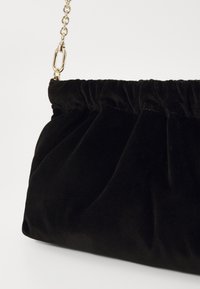 Bolsa de veludo preto com textura franzida, segurada por uma alça de corrente dourada e um fecho, apresentando uma forma suave e arredondada.
