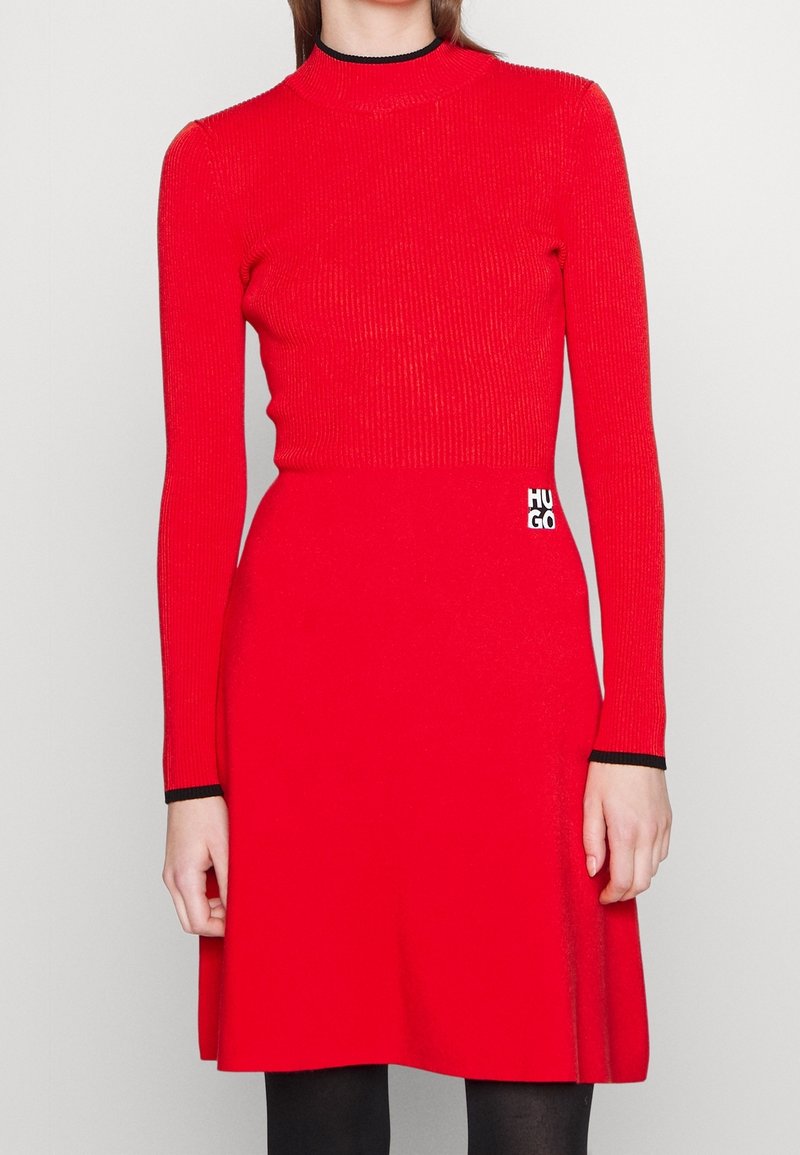 Femme portant une robe rouge ajustée à manches longues avec un liseré noir au col et aux poignets, et un petit logo "HUGO" à la taille.