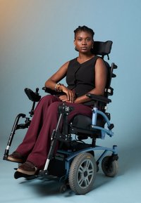 Fauteuil roulant électrique avec un cadre bleu, un siège noir et de grandes roues arrière ; une personne porte un haut noir et un pantalon bordeaux, assise avec confiance.
