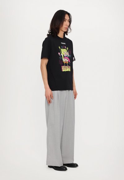 Black cotton t-shirt with a colorful SpongeBob graphic, paired with gray pleated trousers and black shoes. Model stands sideways.