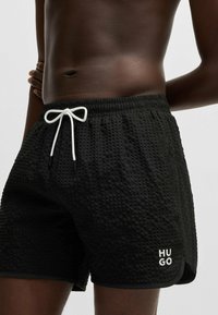 Pantaloni da bagno neri con texture, dotati di una vita con cordoncino e del logo bianco "HUGO" sulla gamba sinistra. Il tessuto presenta un motivo in rilievo.
