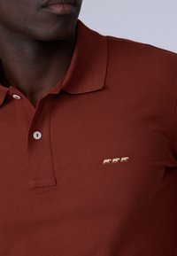 Polo shirt de couleur rouille avec un tissu texturé. Présente un col, deux boutons blancs et un petit design brodé de trois vaches en beige.