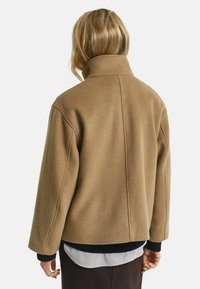 Tan wool-blend jacket with a high collar, dropped shoulders, and a loose fit. Textured fabric with solid color and no visible fastenings.