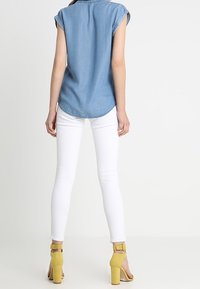 Camicetta in denim azzurro chiaro con maniche arrotolate, abbinata a pantaloni bianchi aderenti e sandali con tacco blocco gialli. Texture liscia e vestibilità casual.