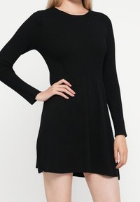 Femme portant une robe noire à manches longues côtelée, avec un haut ajusté et une jupe évasée, posant avec une main sur la hanche.