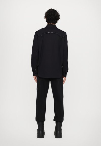 Camisa negra de manga larga con cuello puntiagudo, adornada con tachuelas plateadas en el rendido, combinada con pantalones negros y botas negras voluminosas.