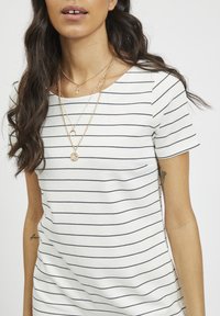 Kobieta w białej koszulce z krótkim rękawem w cienkie czarne poziome paski, z nałożonymi złotymi naszyjnikami i falującymi ciemnymi włosami widocznymi.