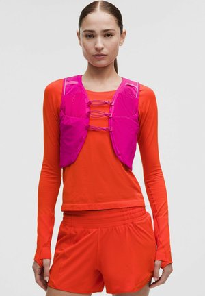 Jonge vrouw die een feloranje sportoutfit draagt met lange mouwen en shorts, gecombineerd met een roze utility-vest met sluitingen aan de voorkant.