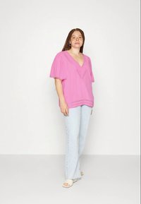Blusa rosa de manga curta com decote em V e detalhes em ilhós, combinada com jeans azul claro e sandálias bege sobre um fundo neutro.