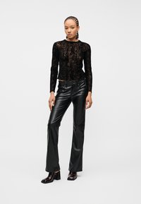 Kvinde stående iført sort mønstret bluse med lange ærmer, sorte læderbukser og mørke ankelstøvler med hæl på hvid baggrund.