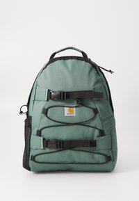 Rucsac verde din țesătură cu curele ajustabile, buzunar lateral din plasă, compartiment frontal cu accesorii negre și o etichetă cu logo. Suprafață texturată.