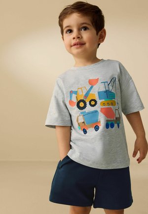 Ung dreng med hånden i lommen iført grå skjorte med farverige illustrationer af byggeplads-køretøjer og marineblå shorts.