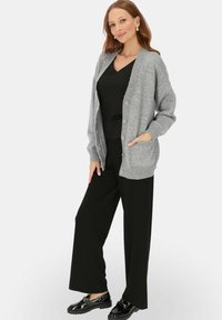 Szary dzianinowy kardigan z prążkowanym kołnierzem i mankietami, z zapięciem na guziki i dwoma przednimi kieszeniami, noszony na czarnej bluzce i spodniach.