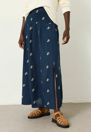 Person mit einem marineblauen, langem Blumenrock mit seitlichem Schlitz, cremefarbenem Pullover und braunen, geschlossenen Sandalen, stehend auf einem grauen Boden.