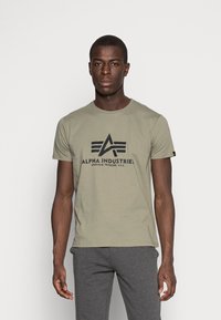 Camiseta de manga corta verde oliva con el logo negro de "Alpha Industries" y acentos geométricos en la parte frontal. La tela parece suave y casual.