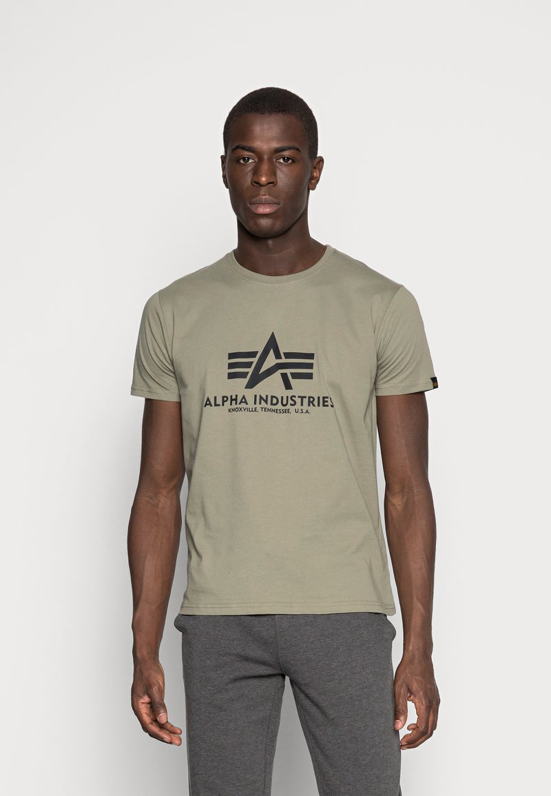 Camiseta de manga corta verde oliva con el logo negro de "Alpha Industries" y acentos geométricos en la parte frontal. La tela parece suave y casual.
