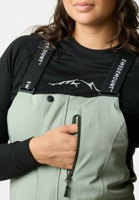 Gröna vadderade overaller med en dragkedjeficka och svarta justerbara remmar, som bärs över en svart ribbad långärmad topp med en bergslogotyp.