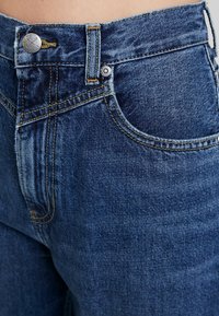 Siniset korkeavyötäröiset farkut, joissa on haalistunut pinta, oranssit saumat, etutaskut ja metallinen napin kiinnitys.