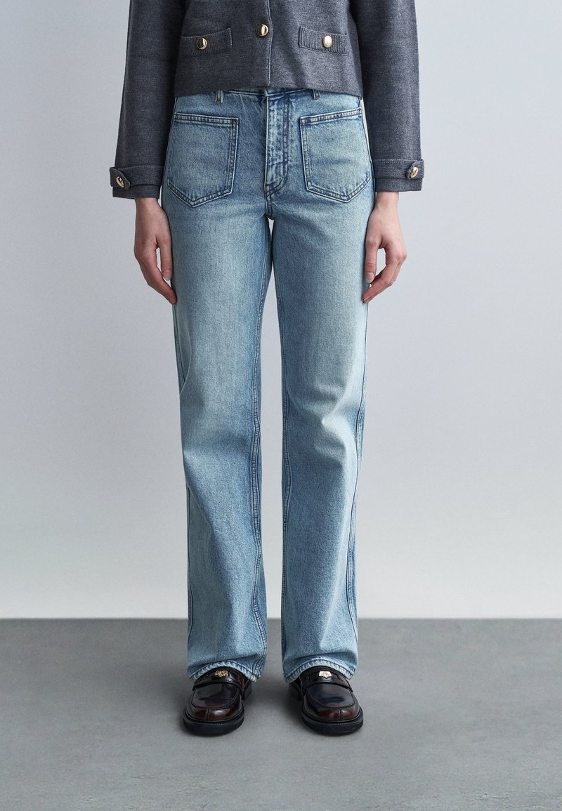 Άτομο που φοράει ανοιχτό μπλε τζιν ίσιας γραμμής, σκούρα μοκασίνια και γκρι μπουφάν με χρυσά κουμπιά, μπροστά σε απλό γκρι φόντο.