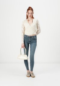 Top blanco de encaje con mangas, jeans azules de cintura alta, zapatillas beige, sosteniendo un pequeño bolso blanco con asas curvas.
