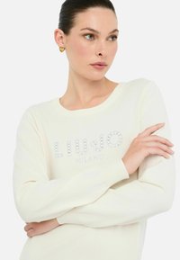 Kreemivärvi kudumipullover, millel on paadi kaelus ja rhinestone'idega kaunistatud logo "LIU JO MILANO" ees. Pehme tekstuur, lõdva istuvusega.