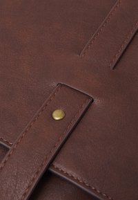 Detail van een bruine leren tas met een messing drukknop, zichtbare stiksels en een soepele textuur met een klepontwerp.