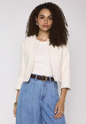 Vrouw met krullend haar, die een witte cropped jas, witte top, high-waisted blauwe jeans, donkere riem en gouden sieraden draagt, staand tegen een effen achtergrond.