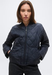 Nuori nainen suorilla ruskeilla hiuksilla, pukeutunut tummansiniseen tikattuun vetoketjutakkiin ja kädet taskuissa, seisoo yksivärisen taustan edessä.