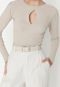 Haut beige à manches longues côtelées, avec une découpe en forme de clé et de légers fronces aux épaules, associé à un pantalon blanc taille haute et une ceinture assortie.
