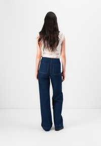 Femme aux longs cheveux foncés, portant un haut blanc en dentelle sans manches et un jean bleu foncé taille haute à jambes larges, debout de dos.