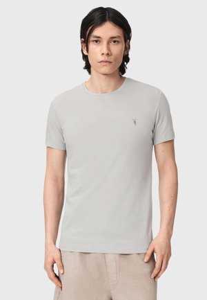Jeune homme aux cheveux foncés mi-longs portant un T-shirt gris clair à manches courtes et un pantalon beige, debout devant un fond uni.