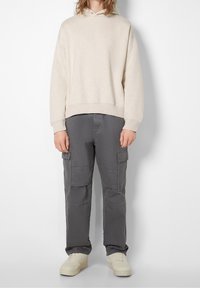 Beige bomullshuvtröja med dragsnöre i huvan, kombinerad med grå cargo-byxor med flera fickor; vita chunky sneakers avslutar looken.