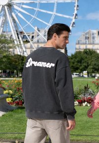 Homme en sweat-shirt sombre avec le texte « Dreamer » dans le dos, debout dans un parc près d'une grande roue et de fleurs par une journée ensoleillée.