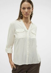 Chemise blanche à boutons avec des rayures verticales, dotée de deux poches poitrine, d'un col en V et de manches retroussées. Texture douce et tissu léger.
