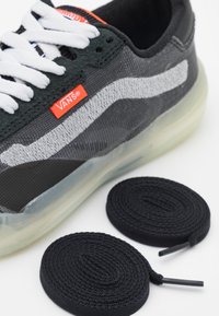 Sapatilhas Vans em preto e cinza com atacadores brancos, sola de borracha e a faixa lateral característica. Inclui dois atacadores pretos em rolos circulares.
