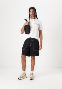 Svarte cargo shorts med hvit søm, en hvit t-skjorte med gul tekst, og beige sneakers. En liten svart crossover-veske bæres.