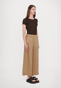 Hnědý pletený top, široké béžové kalhoty s elastickým pasem a černé platformové sandály. Modelka stojí rovně proti bílému pozadí.