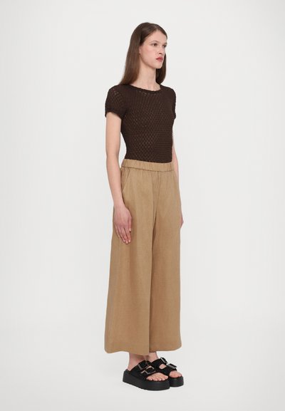 Brązowy sweter, szerokie beżowe spodnie z elastyczną talią oraz czarne sandały na platformie. Modelka stoi prosto na białym tle.