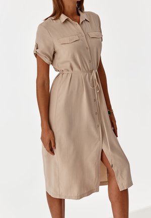 Femme portant une robe beige à manches courtes boutonnée avec poches poitrine et ceinture nouée à la taille, debout sur un fond blanc.
