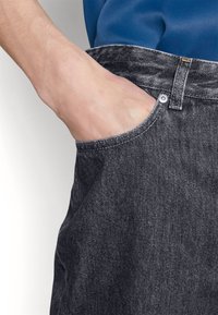 Vaqueros de denim negro con un bolsillo frontal, que presentan un botón metálico visible y un detalle de costura. La tela tiene un acabado ligeramente texturizado.